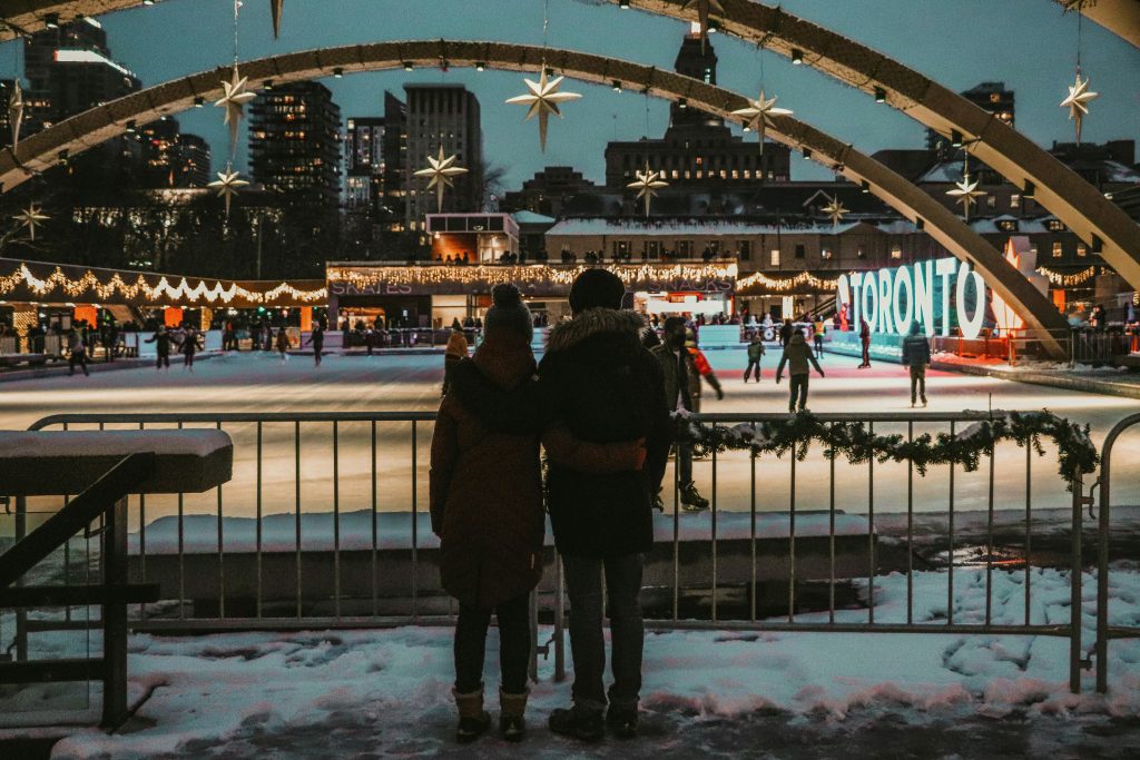 Couple embraces in winter attire overlooking ice rink in festive Toronto setting.