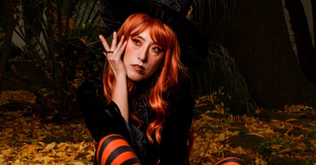 Young woman in witch costume sits among autumn leaves outdoors.