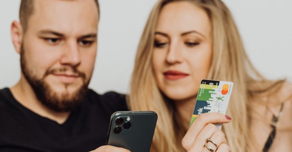 A bearded man and woman shopping online using a smartphone and credit card, sitting indoors.