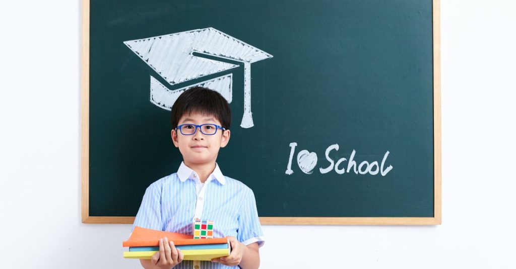 A smiling young boy stands in front of a chalkboard with a graduation cap drawing and "I love school" text.