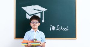 A smiling young boy stands in front of a chalkboard with a graduation cap drawing and "I love school" text.