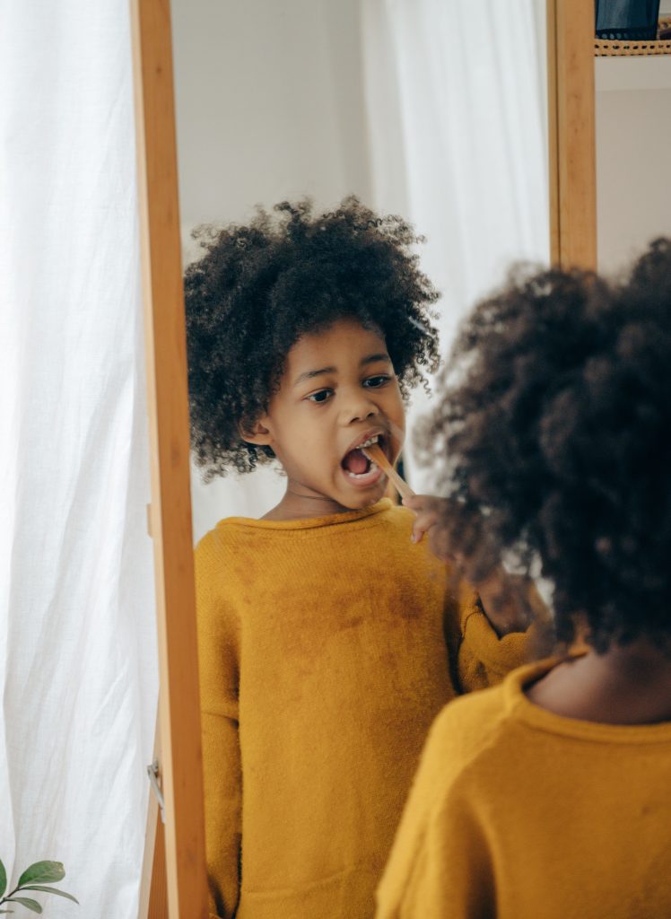 Ethnic child in yellow sweater using toothbrush for cleaning teeth while standing in bathroom looking at mirror