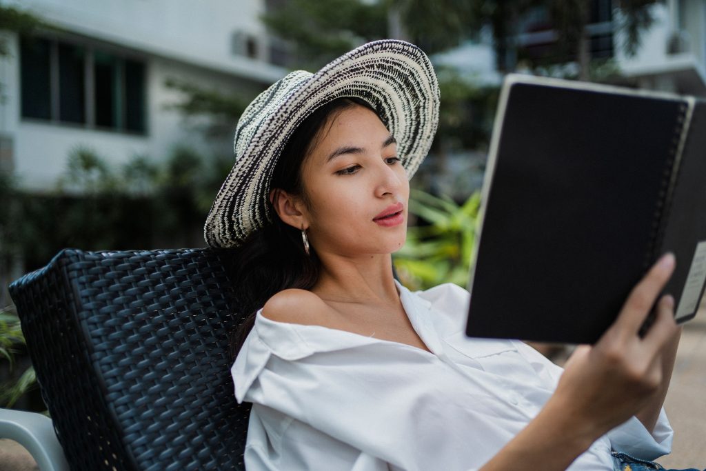 Focused young ethnic lady in white shirt and straw hat lying on sunbed in hotel yard and reading information in notebook