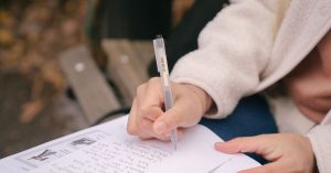 Crop unrecognizable woman in casual outfit sitting on wooden bench while taking notes on paper near textbook in street with foliage on ground in daylight in autumn