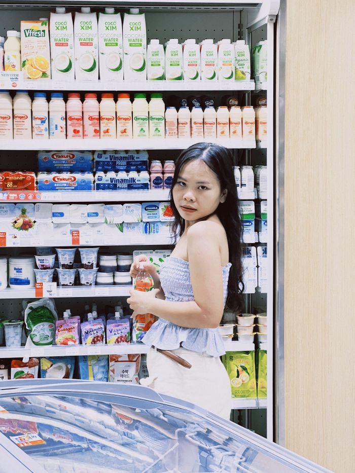 A woman holding a bottle stands in the dairy section of a supermarket, surrounded by various products.
