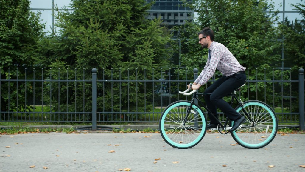 Professional man in office attire rides bicycle on a city pathway lined with greenery.