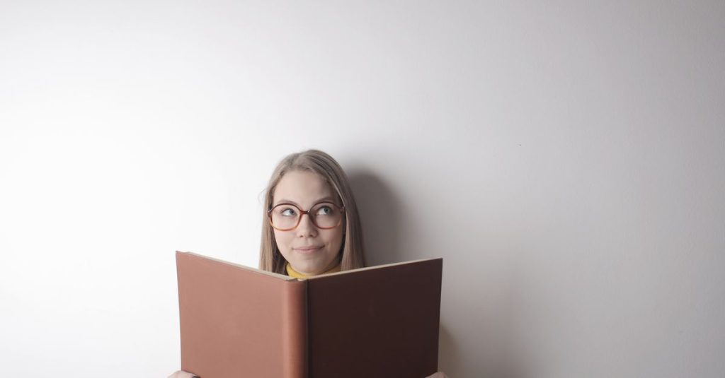 Positive young curious female wearing eyeglasses looking up while standing against gray wall with big opened book with brown cover in hands