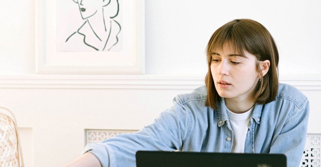 A young woman working diligently in her home office, reaching for stationery while focused on a laptop.