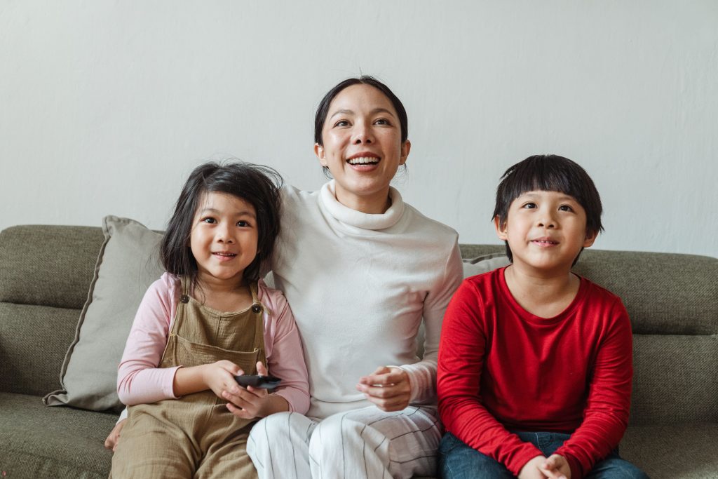 Excited ethnic mother and little son and daughter in casual clothes watching interesting movie on TV while having rest in living room together