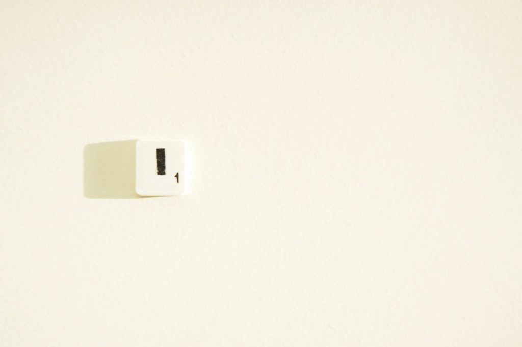 A white Scrabble tile with the letter 'I' and number '1' with a shadow on a light wall.
