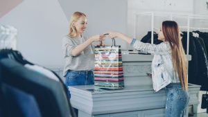 Two women interacting at a retail store counter, exchanging a colorful shopping bag.