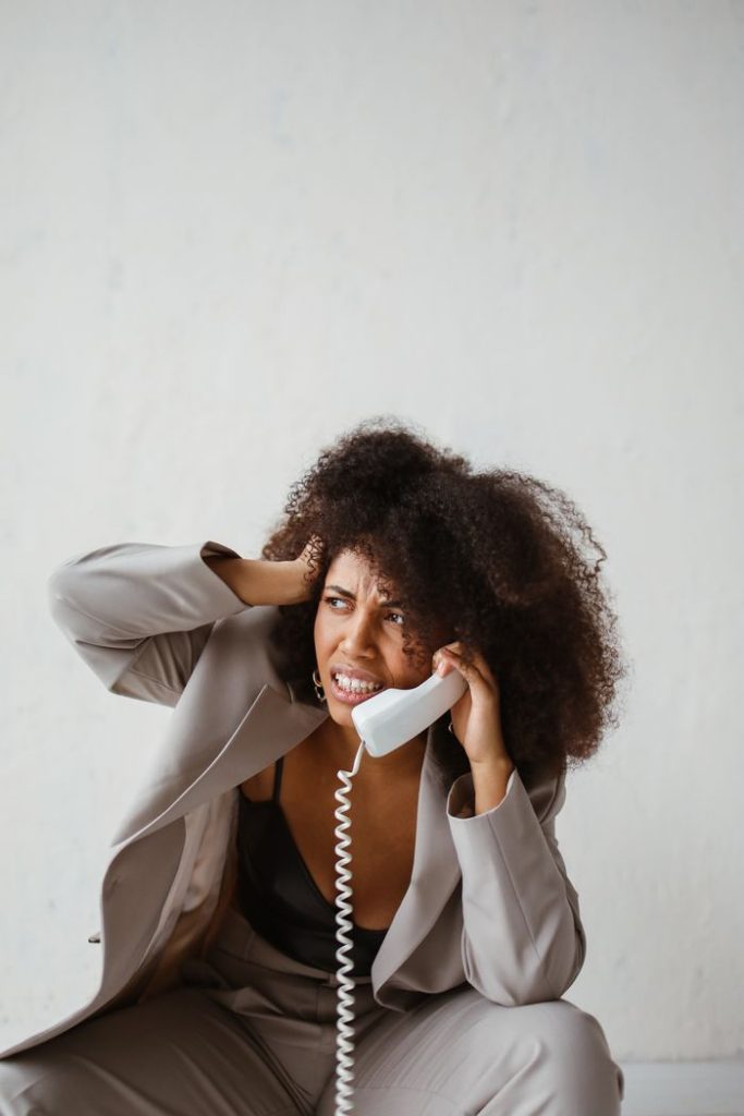 Stressed African American woman with afro hairstyle sitting and holding a phone to her ear indoors.