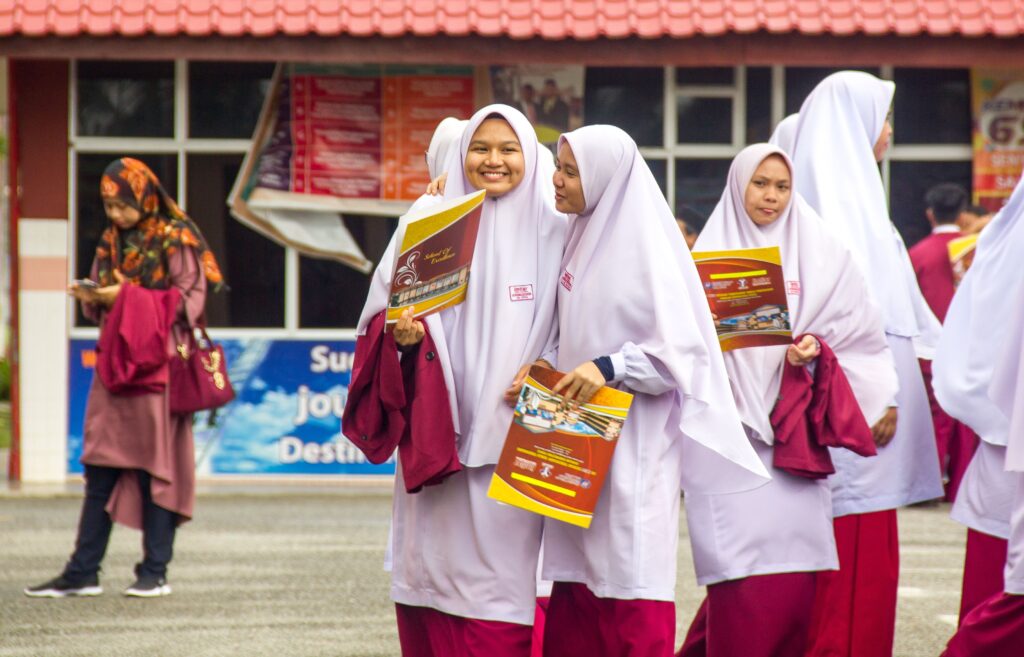 A joyful group of schoolgirls in white hijabs and maroon uniforms smiling outdoors, holding brochures.
