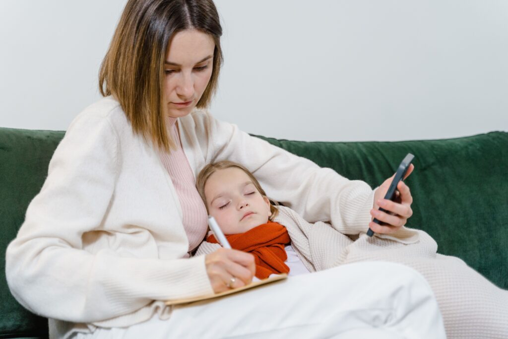 A mother comforts her sick child while writing notes and using a smartphone, portraying modern caregiving.